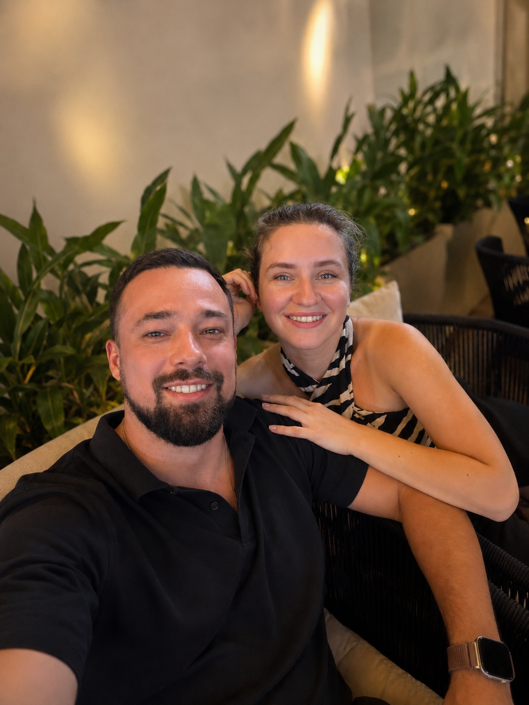 A smiling Roman Nekrasov and Diana Nekrasova posing together in a cozy indoor setting with green plants in the background.  Roman in the foreground wears a black polo shirt, and Diana behind him wears a black and beige patterned top.